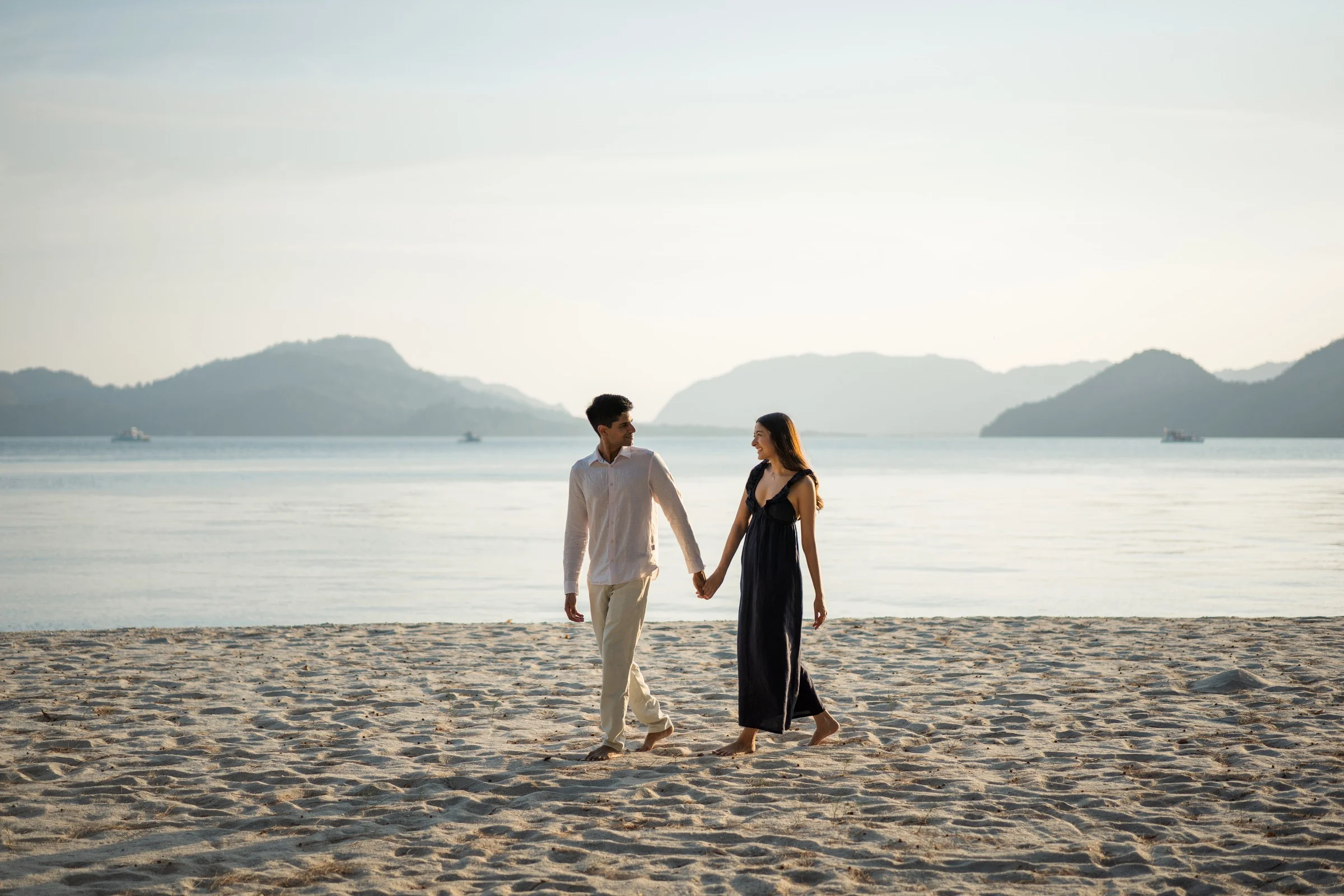 Alisha and Kunal walking hand in hand on the beach, with the lake and mountains behind them