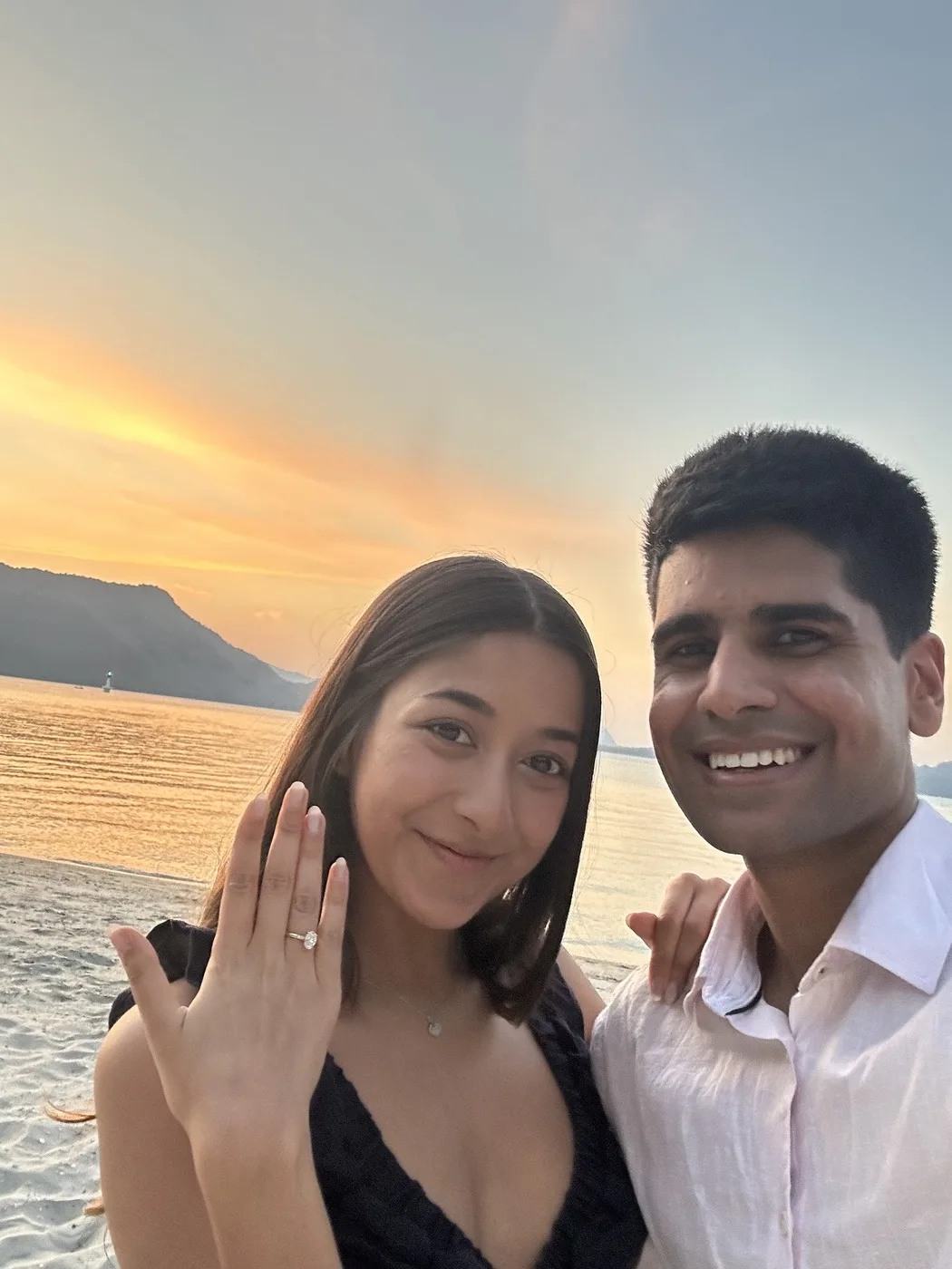 Alisha and Kunal taking a selfie beside the lake at sunset, showing their rings